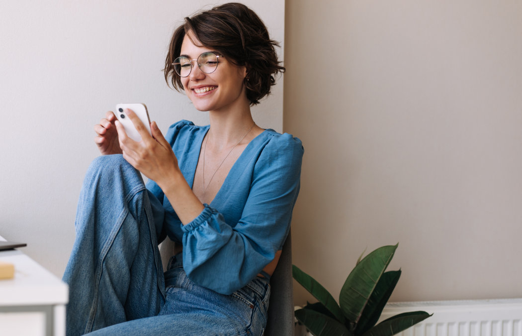 Woman in a blue blouse sitting against a wall, smiling at her phone. A potted plant is visible beside her.